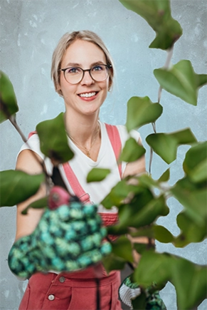 A woman in a gardener's outfit is holding a plant up to the camera